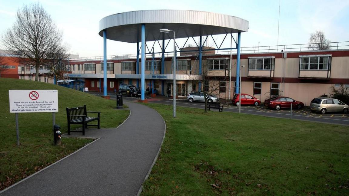 A two-storey building with the words 'main entrance' written above a door. A semi-circular overhang with six pillars is in front of it. In front of that are several cars and a single carriage road. A winding path with grass and a bench is in the foreground.