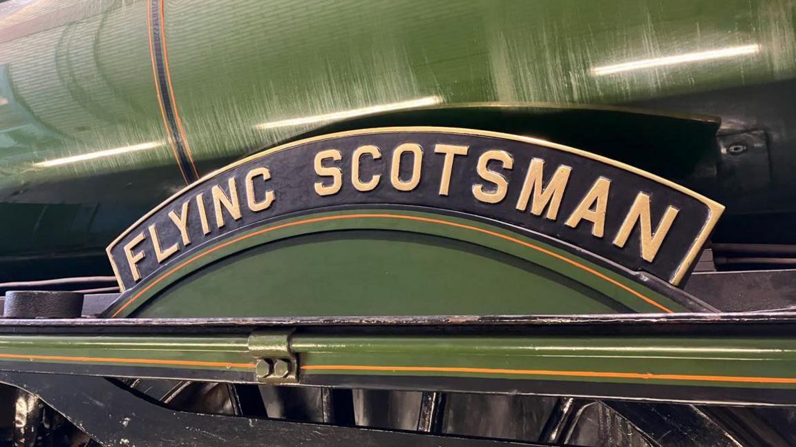 A green-painted metal cylinder with a curved black sign which has the words flying Scotsman written on it in gold lettering and some black-painted metalwork below