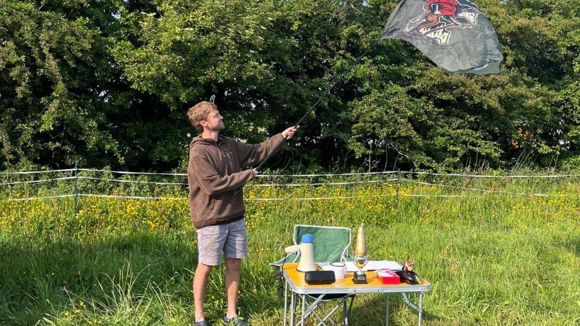 Mr Weston wearing a brown fluffy jumper and stone coloured shorts with dark shoes. He is flying a kite branded with a wolf on it and standing on lush grass with trees in the background. There is a pop up camping table with a trophy, flask and other bits on, and a green camping chair behind it.