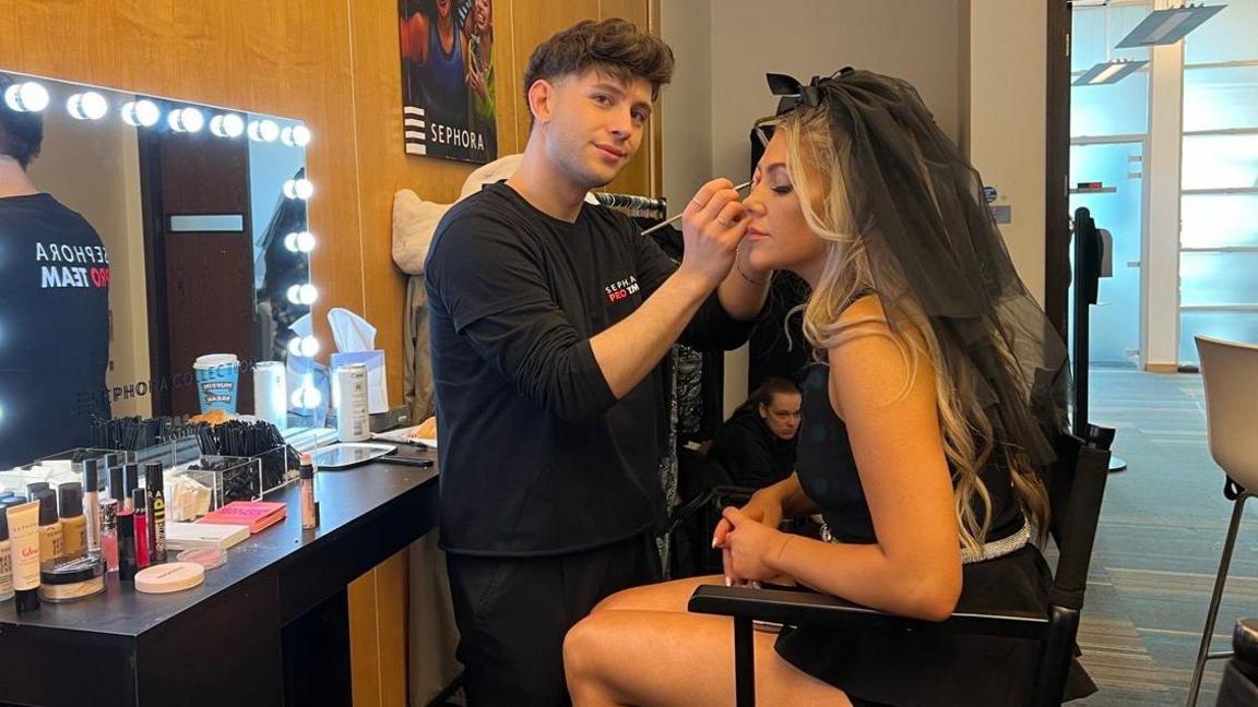 A man has short brown hair and is wearing a black t-shirt. He is standing next to a makeup vanity that is lit up with bright lights. He is putting eye make-up on a woman with long blonde hair. She is wearing a black dress and a black headpiece 