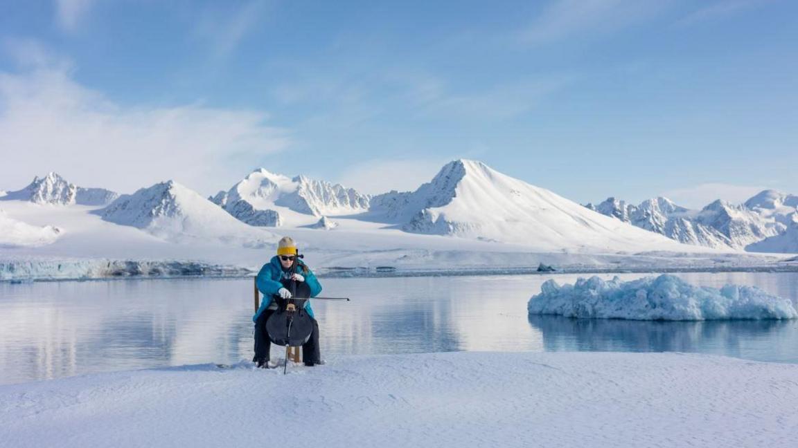 A woman wearing a blue coat, sunglasses and a yellow hat sits on a chair on ice in front of a lake, in the background are snowy mountains. She is playing a cello.