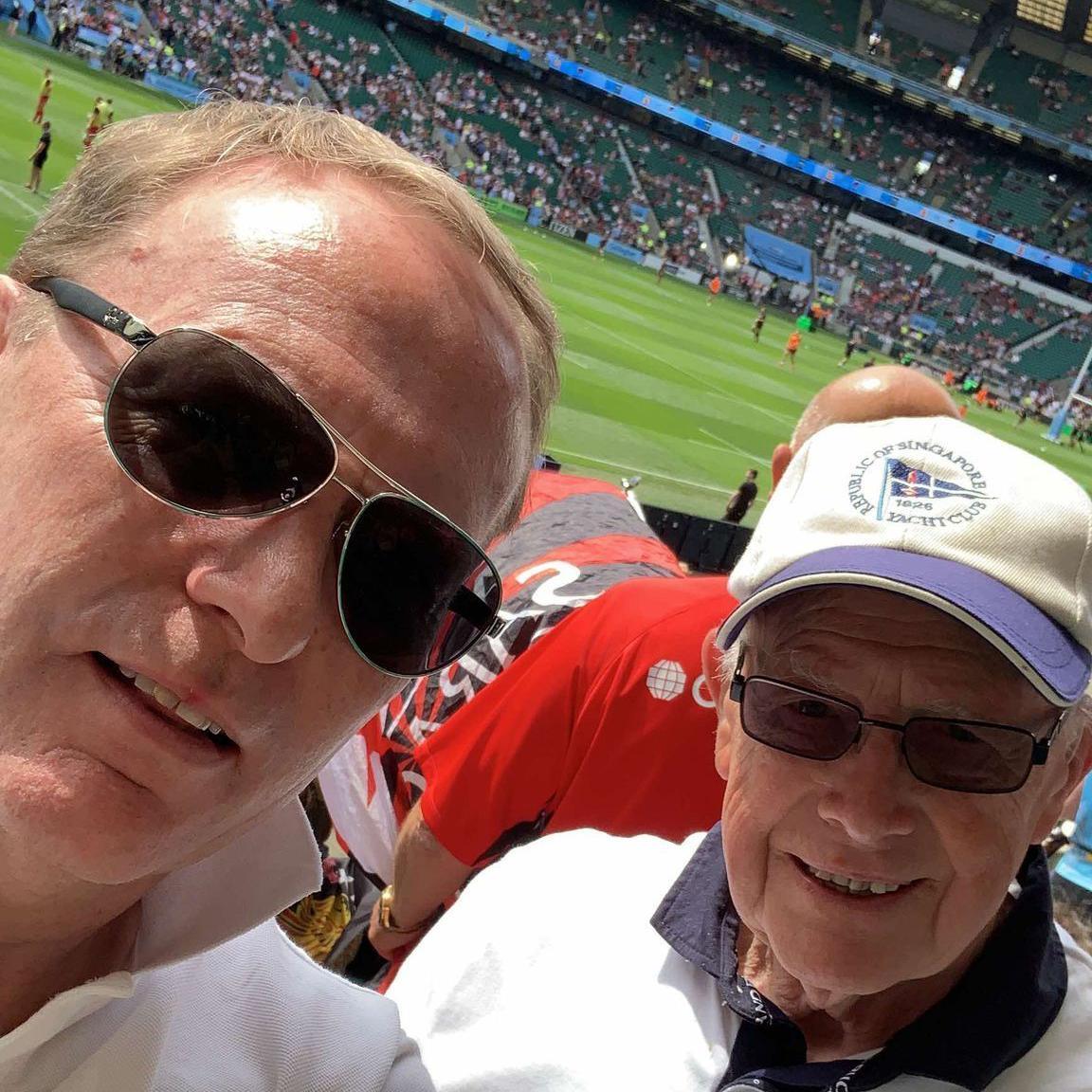 A father and son smiling from the stands of a rugby stadium ahead of kick off