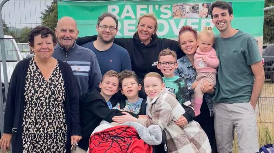 A photo of a group of men and women, with including four children and a baby. They all have their aims around each other and are smiling. Behind them is a banner saying '"Rafi's recovery".