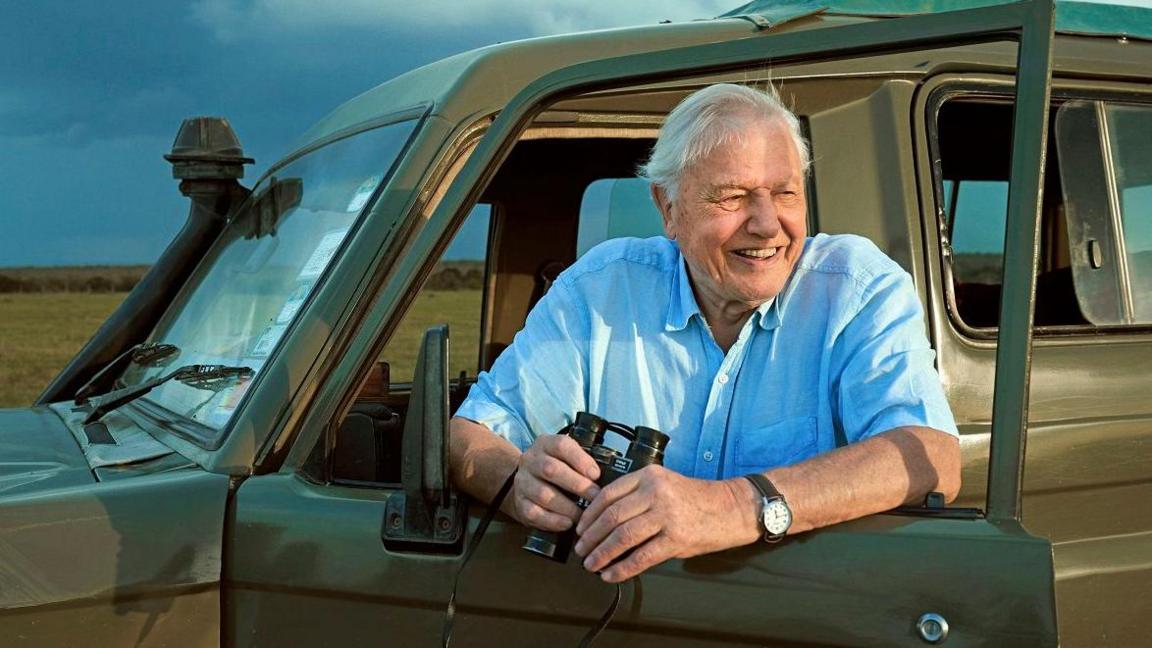 David Attenborough standing in the open door of a jeep holding a pair of binoculars