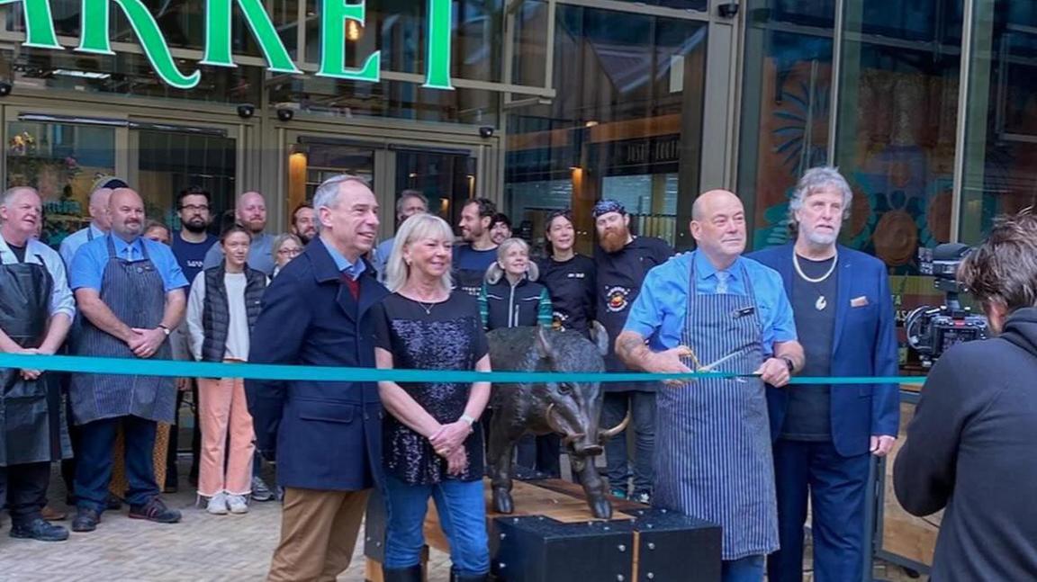 Geoff Hughes in blue apron cuts a blue ribbon at the market opening ceremony.