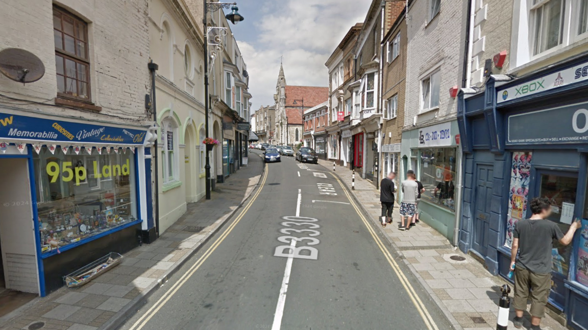 A Google street view screenshot of a narrow high street with shops on either side. There's a church at the end of the road and it's a sunny day.