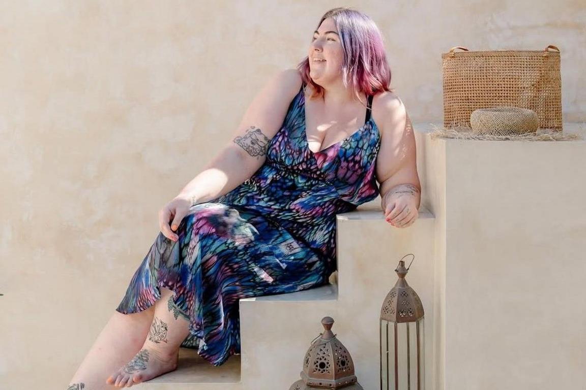 A woman in her early 30s sitting on some steps in the sunshine. She is looking away from the camera and wearing a long pink and blue summer dress, there is a pinkish bare plaster wall behind her and some Morrocan style lamps in the foreground. A basket weave bag sits on the step above her.