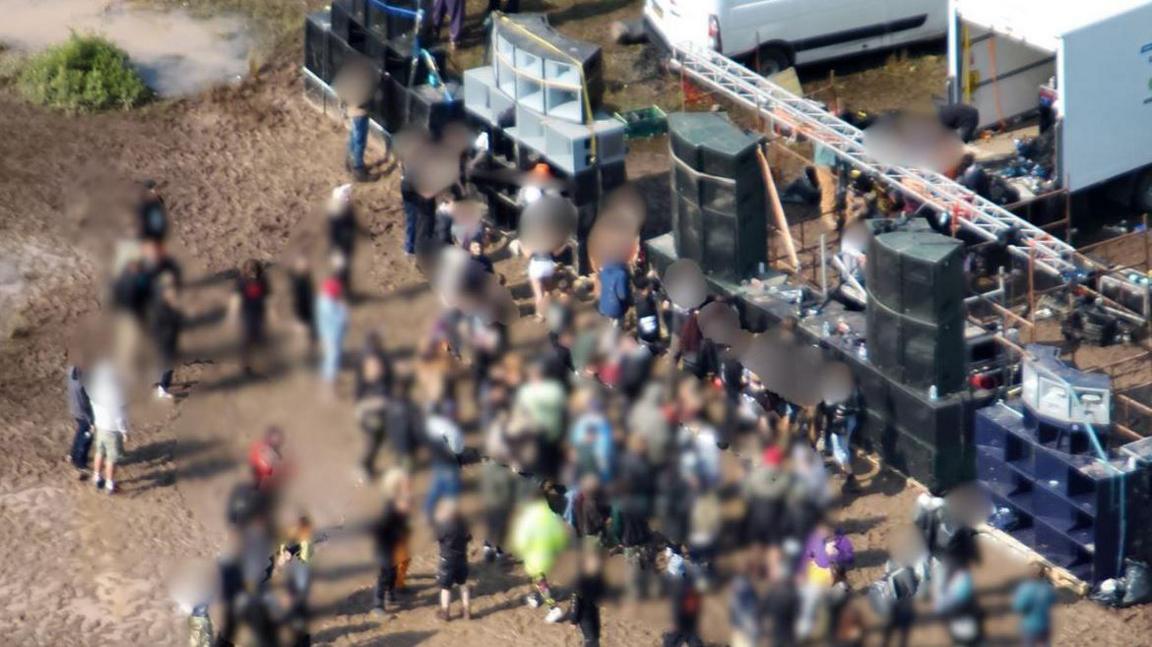 A crowd of about 60-70 people gathered around a makeshift stage made of stacked speakers. The ground is dusty and dirty, and everyone has their faces blurred out. There are large white vans parked in the background.