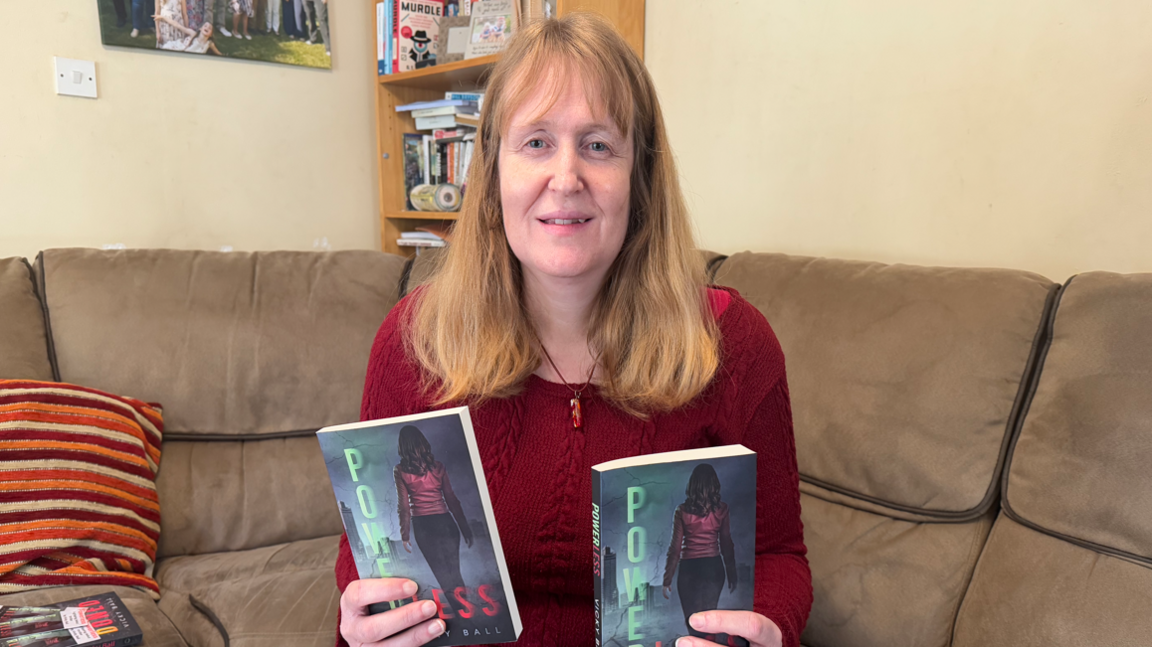 A woman with shoulder-length hair sitting on a brown sofa while holding two copies of her book Powerless. She is looking into the camera
