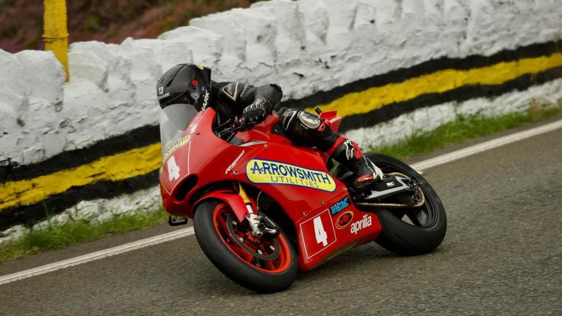 Lewis Arrowsmith wearing black leathers and helmet on his red Supertwin machine, which has the number four on the front. he leads over as he passed a stone wall that is painted white.