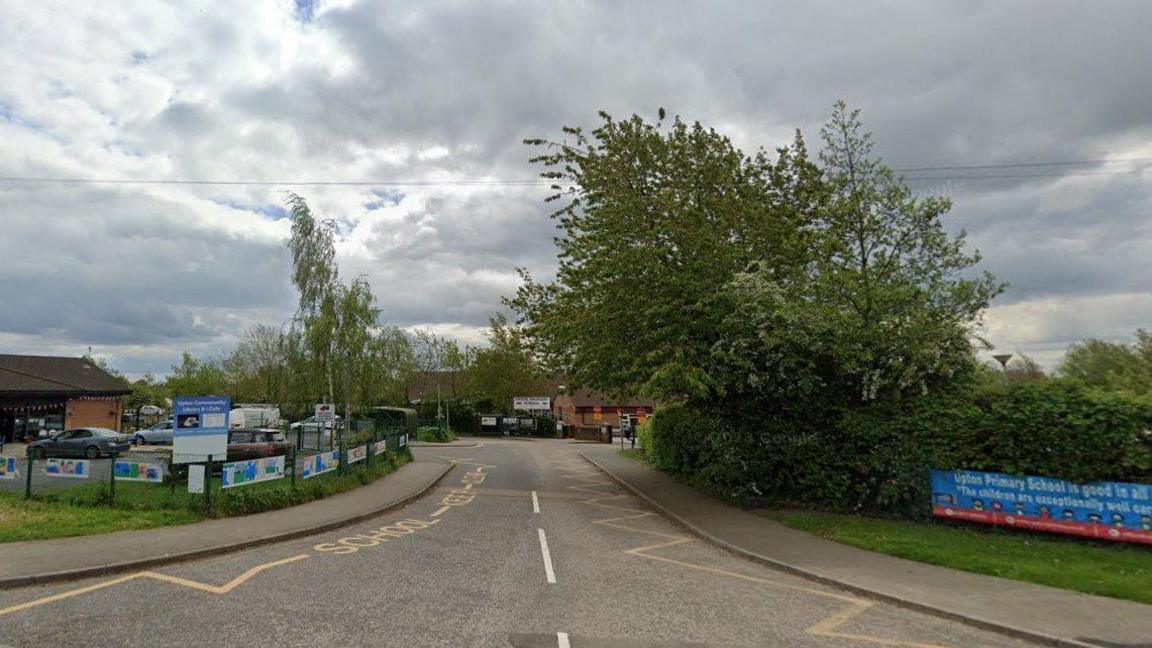 The entrance to a Upton Primary School. There are colourful posters on the approach to the school with trees and greenery surrounding the site. 