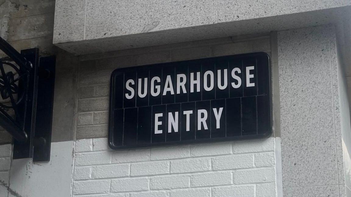 A black sign on a white brick wall that says, sugarhouse entry