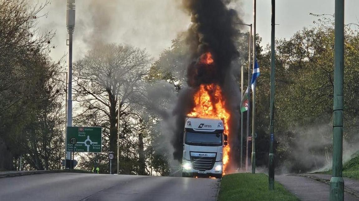 A photo taken from a distance of a lorry on fire on the side of a road. Flames can be seen coming from all sides of the vehicle and thick black smoke billows up into the sky. A pavement and grass runs alongside the road and there is a green roundabout road sign on the left. 