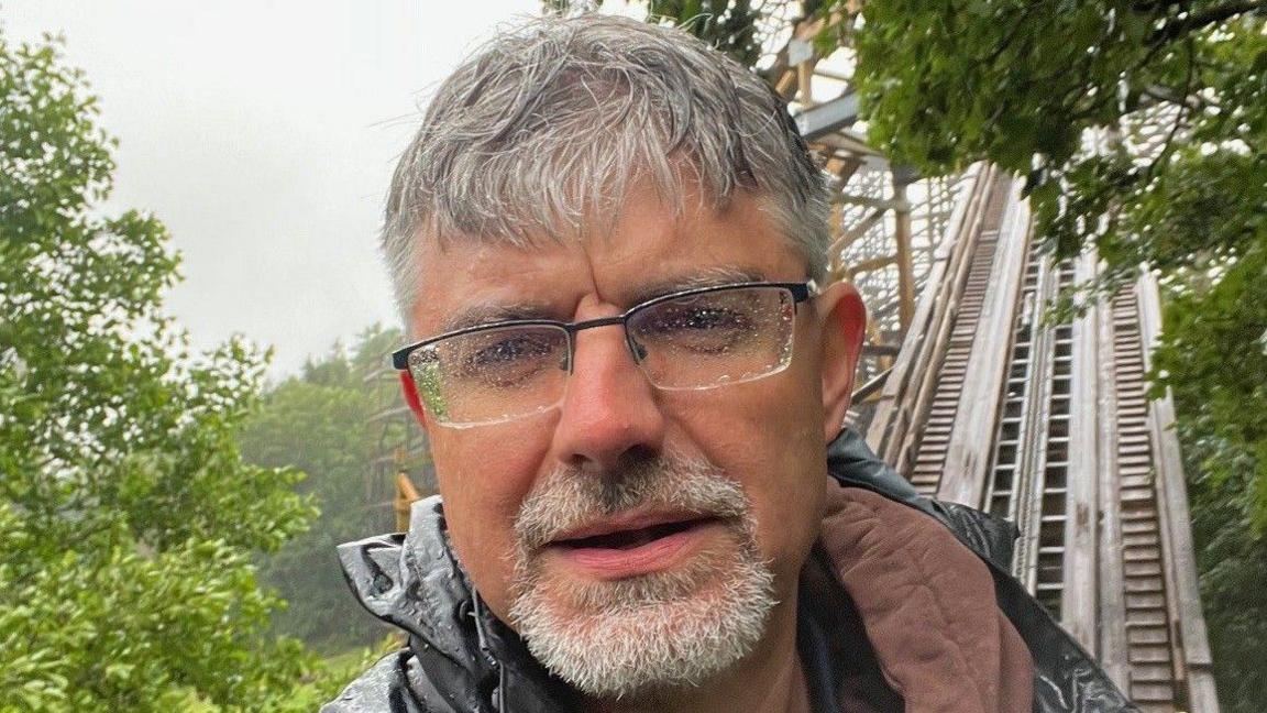 Korey Kiepert standing on the Megafobia ride on a wet day, he has short, grey hair and a goatee. There are raindrops on the lenses of his glasses and the track can be seen behind him. Trees can be seen on either side of Korey. 