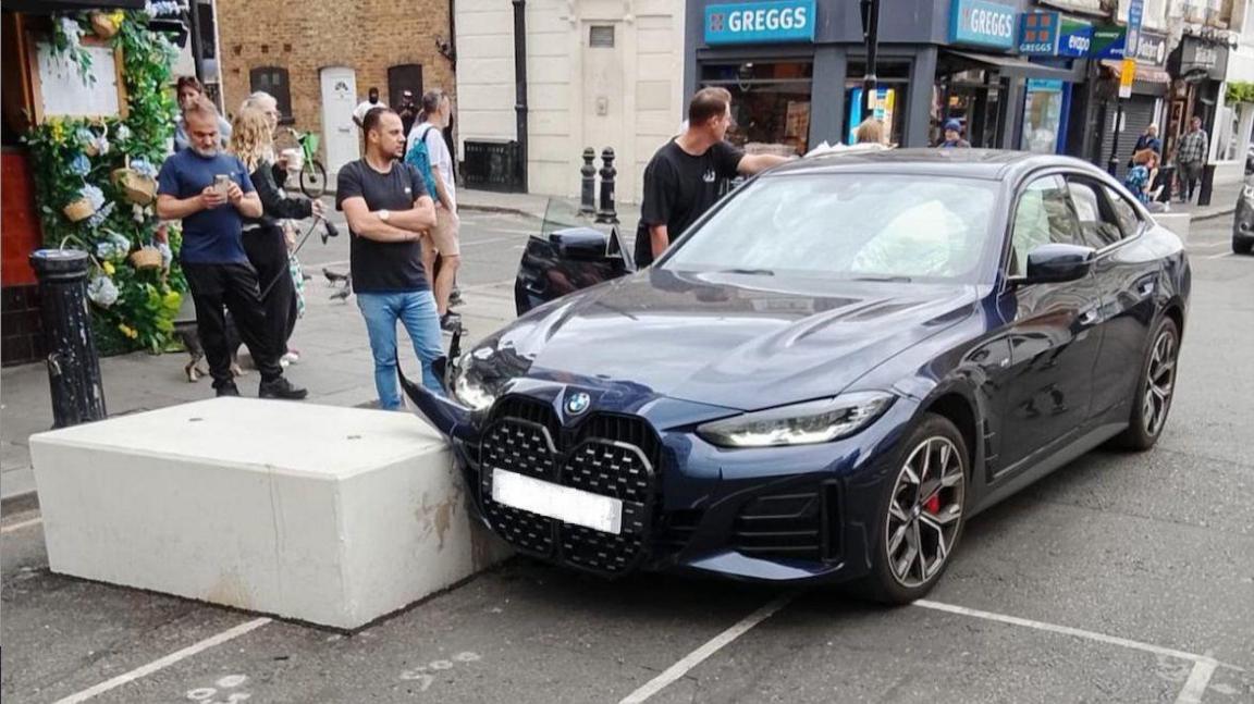 A dark blue BMW car crashed into a low, light grey concrete block. The car has hit the block in its front right corner, with damage to the bodywork on that part of the car. A few men have gathered around, with one leaning on the side of the car, and another next to the car with crossed arms. The driver's side door is open. The street is visible in the background, with a pavement running alongside the road where the crash has happened, and a Greggs bakery behind the car.