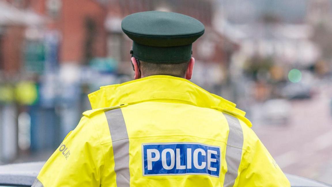 A police officer with short hair stands with his back to the camera. He is wearing a hi-vis coat with 'police' on the back and a green hat. The background is blurred.