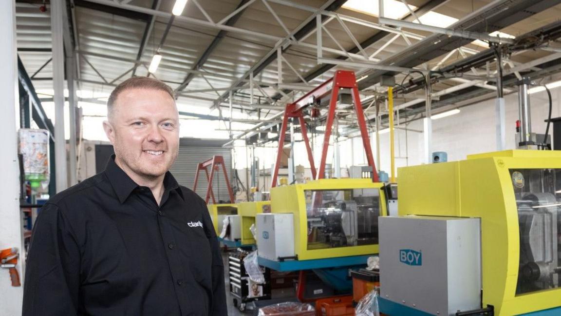 Head and shoulders of a man, smiling, wearing a black shirt, standing in a factory with a row of yellow manufacturing units in the background