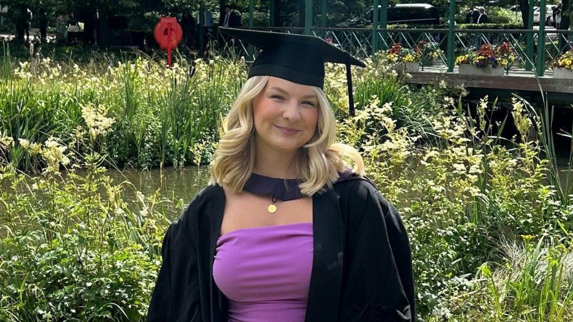 A blonde woman wearing a graduation cap and gown smiles as she stands by a pond in a park