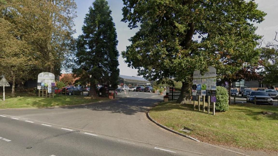 An entrance into a garden centre. There are tall green trees on both sides of the road and two signs in front that read "Bell Plantation".
