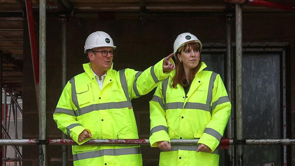 Sir Keir Starmer and Angela Rayner on a building site in Cambridgeshire in 2024. They are both wearing hard hats and high vis jackets, and are framed within scaffolding poles.