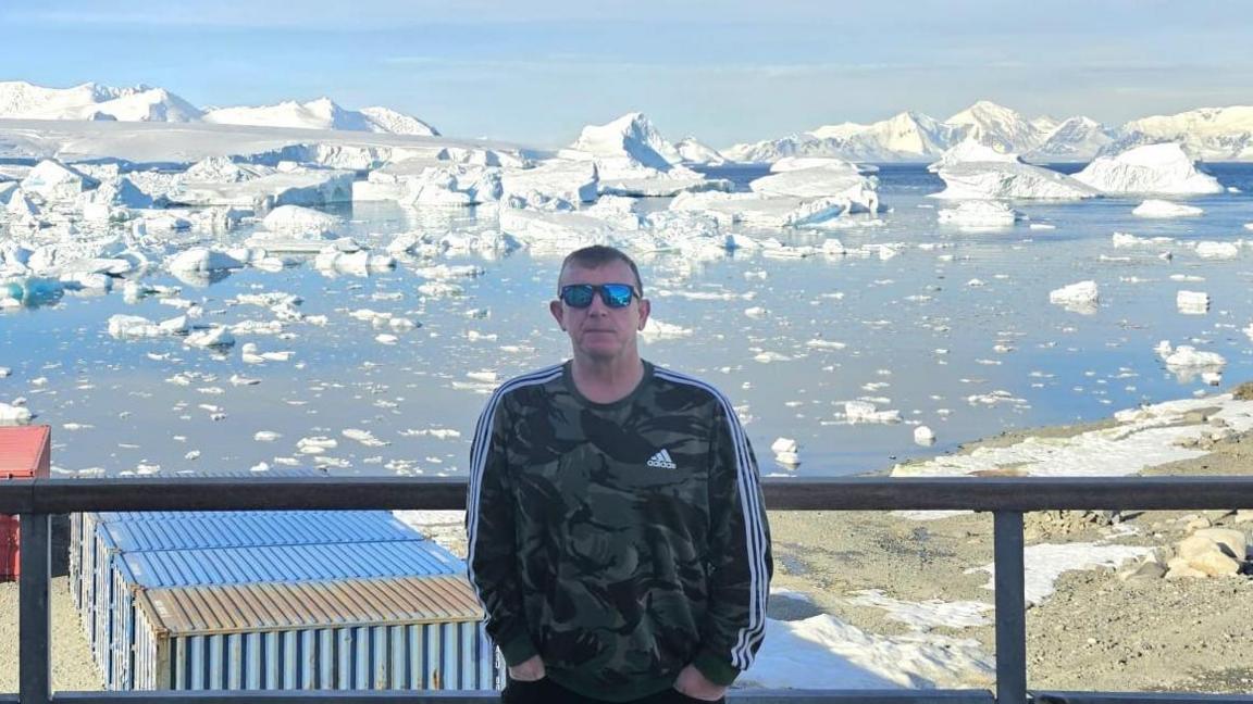 Michael Young wearing sunglasses and a camouflage Adidas long-sleeved top, with his hands in his pockets, leaning against a metal fence. Behind him are blocks of ice floating in water beyond a series of shipping containers.