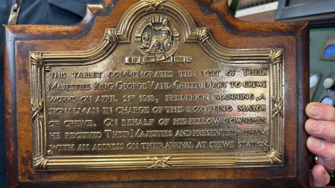 A brass plaque on a wooden background words written on it. The words start with "This tablet commemorates the visit of their majesties King George V and Queen Mary" and goes on to say when the visit was and where. The plaque is on a wooden background and a hand can be seen on one side holding it.