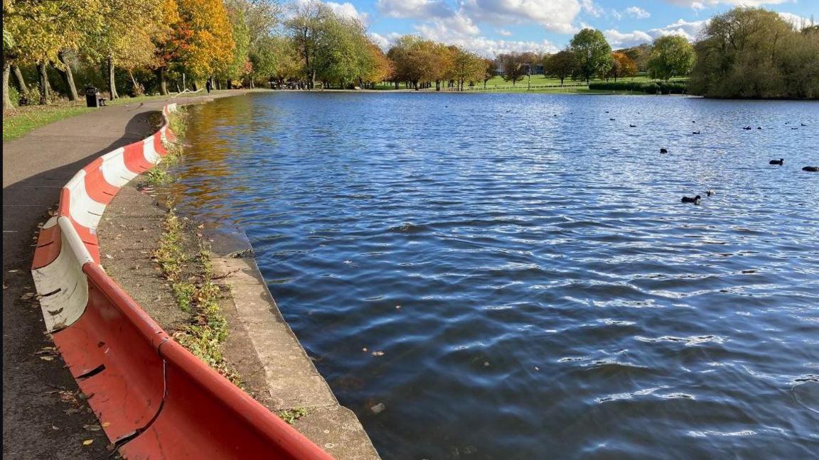 Lake at Pontefract Park with ducks on the water, autumn trees and grassy bank to the left edge, blue sky with white fluffy clouds, red and white heavy plastic safety barriers around the perimeter.