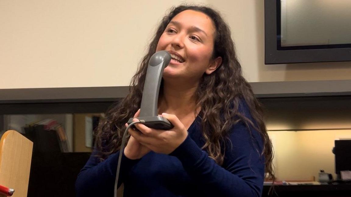 Lara Harris smiling and singing into a grey intercom. She has long, wavy brown hair and is wearing a navy jumper.