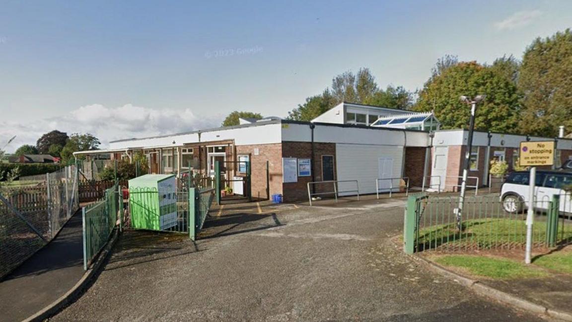 The gates are open to the entrance to Much Birch Church of England Primary School, which is a single storey building. A car is parked to one side and there is a No Stopping sign.