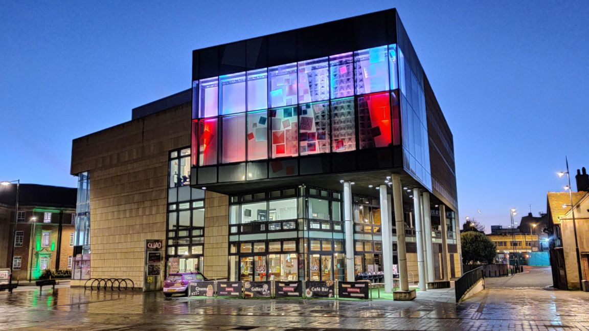 The Quad building in Derby, which has light coloured bricks on the left and four floors of glass windows on the right - the top two rows of glass windows are coloured blue and red, with shapes on. It is dark outside. 