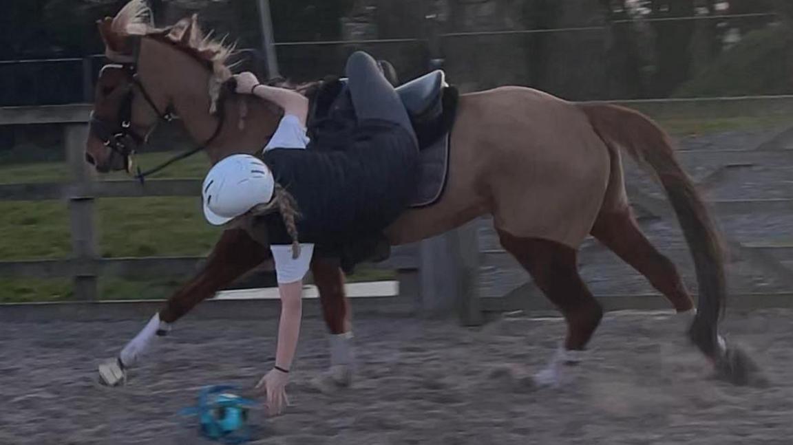 A young woman hanging from a horse's back to the floor to pick up a ball. She has a long blonde plait and is wearing black jodhpurs, a black T-shirt with white sleeves and a black helmet. The horse is chestnut with white socks and is cantering around a sandy ring.