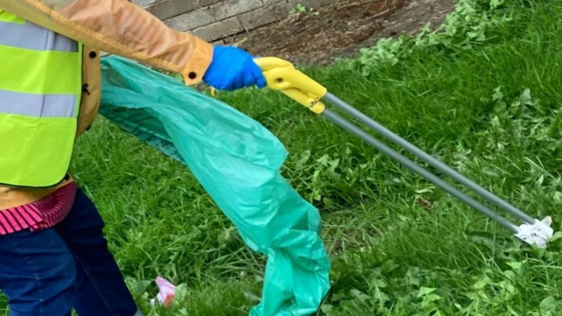 Partial view of a person holding a litter picker, grasping a bit of paper, in their right hand and a turquoise bin bag in the right against a grassy background