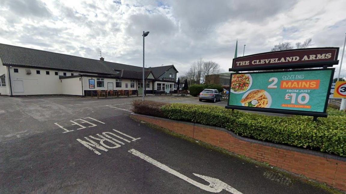 A sign with the words The Cleveland Arms is on the right hand side of the photo. A building goes from the left of the image to the middle, where a car is parked.