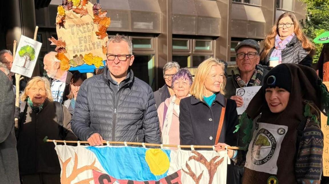 A group of protestors carrying banners and some wearing tree costumes stand together ahead of a council meeting