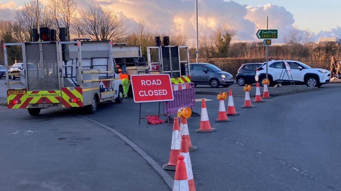 A red 'road closed' sign by a roundabout. Four orange cones are in front of the sign and a highways vehicles is behind it. Cars are queuing at the roundabout on the opposite carriageway and a van is travelling around the roundabout