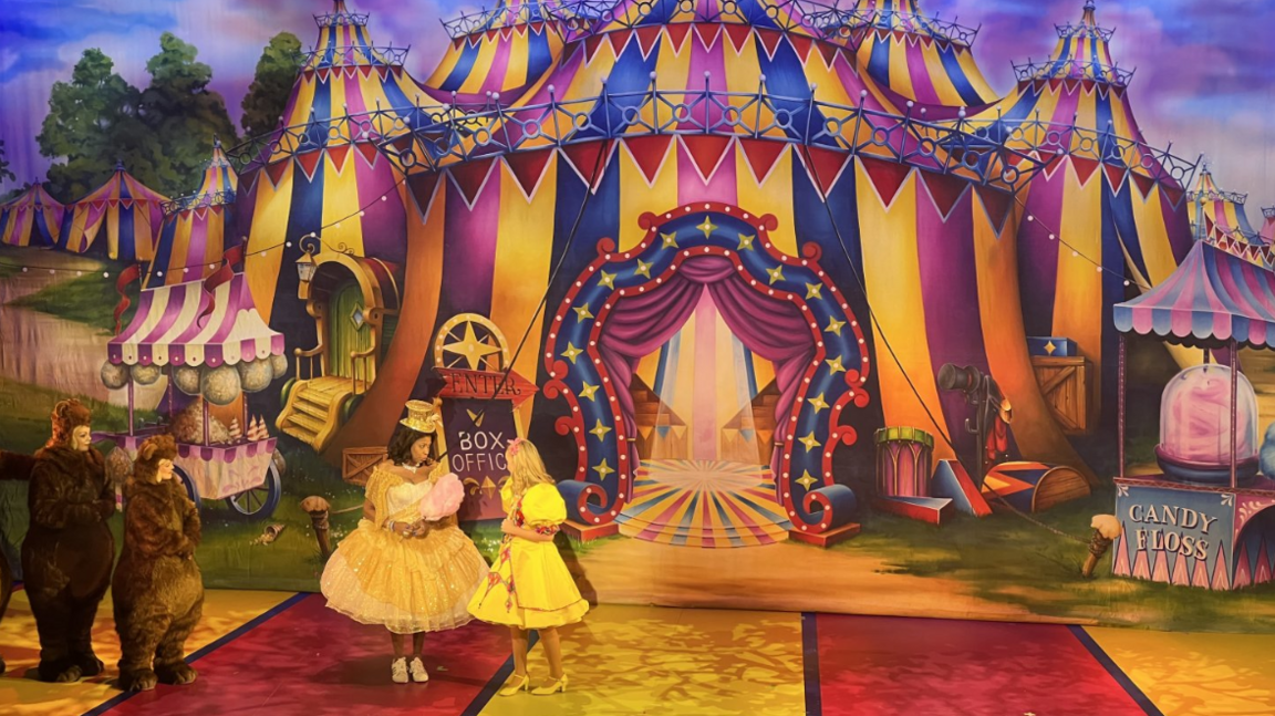 Cast members in Goldilocks and the three bears on stage at the Bristol Hippodrome. Two bears are visible in brown bear costumes and goldilocks is wearing a yellow dress. Another character - Candy Floss - is wearing a gold dress. There is a marquee stage set behind them of yellow, red, blue and pink. And a candy floss stand to the right.