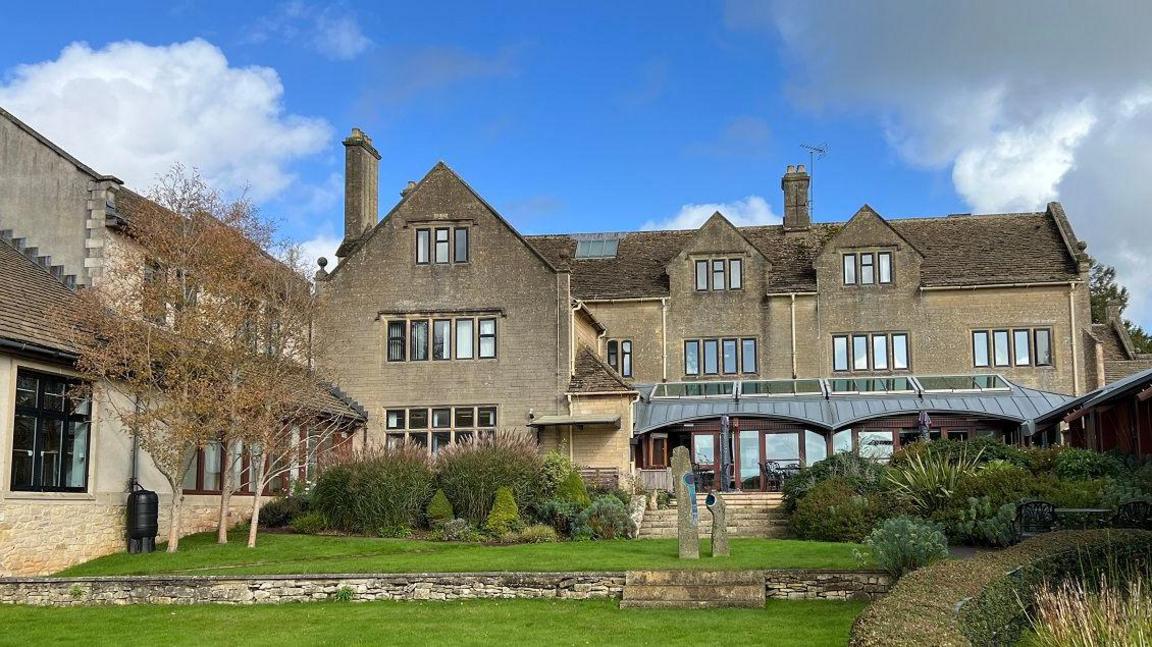 A large three storey building with Cotswold stone, with a grassy lawn in front of it. 