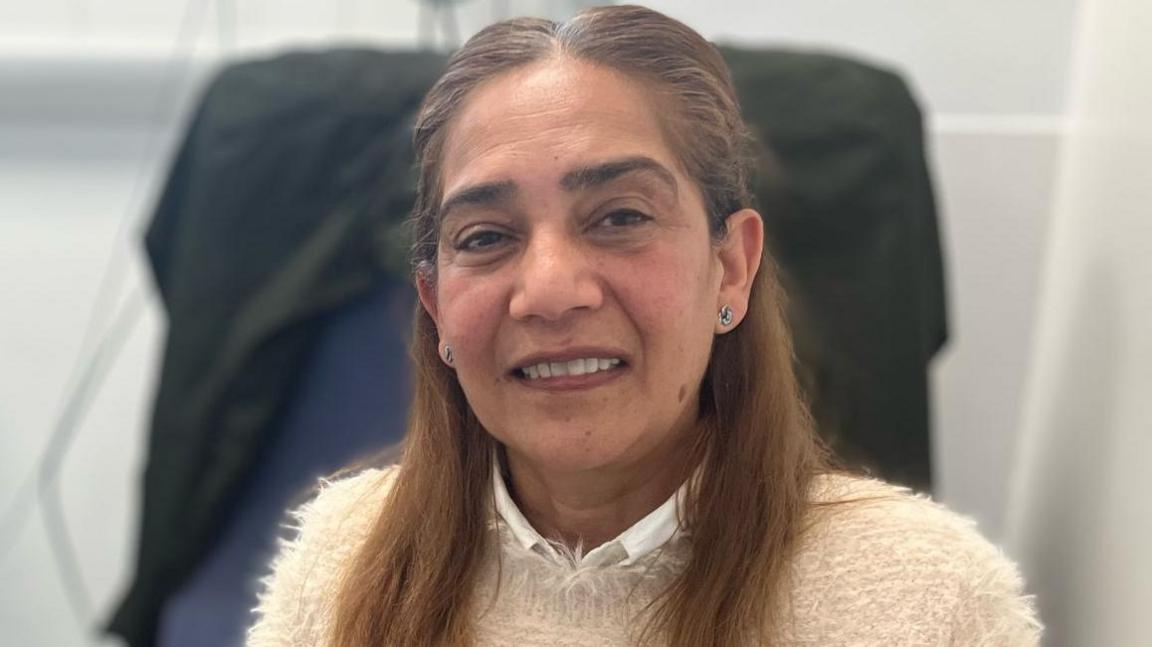 Nafisabanu Ahmed at the endoscopy centre at Leicester General Hospital. She has long brown hair and is looking at the camera. There is a chair behind her that has a black jacket.