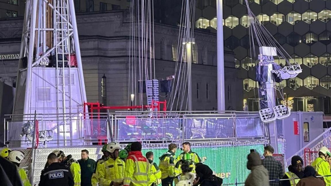 A large fairground ride with what appears to be tangled swings surrounded by emergency services.