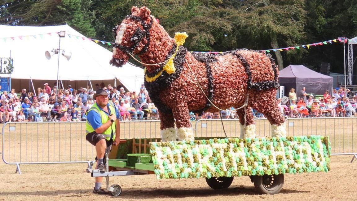 A man wearing shorts, a blue shirt, a black cowboy hat and a yellow high-vis jacket stands next to a large horse made out of flowers for a display at the North Show in Guernsey. A large crowd is watching on.