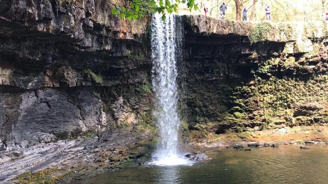 A waterfall plunges into a large pool of rocks, with several people at the top