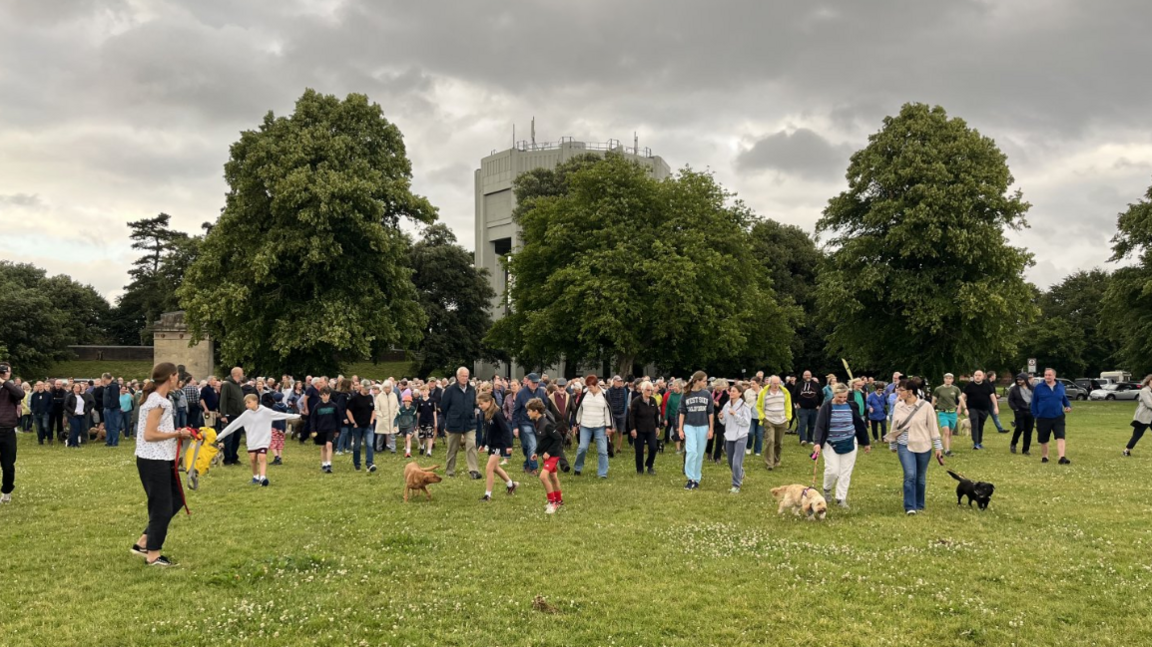 A large group of people including children and dogs walk in a group near the water tower on the downs in Bristol