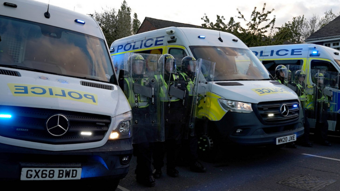 Police officers with riot shields stand in a line next to police vans.