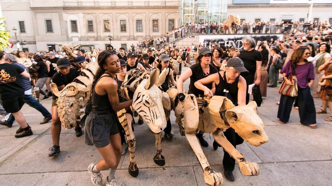 Crowds on the street watch life-sized cardboard animals being processed around a public square