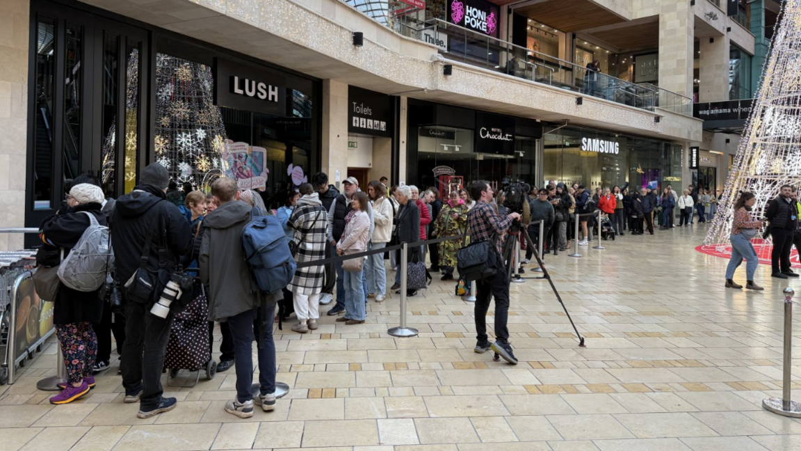 A long queue of people wait to enter a newly opened M&S store in Bristol's Cabot Circus shopping centre, there is a tall white artificial Christmas tree decoration in the right hand side