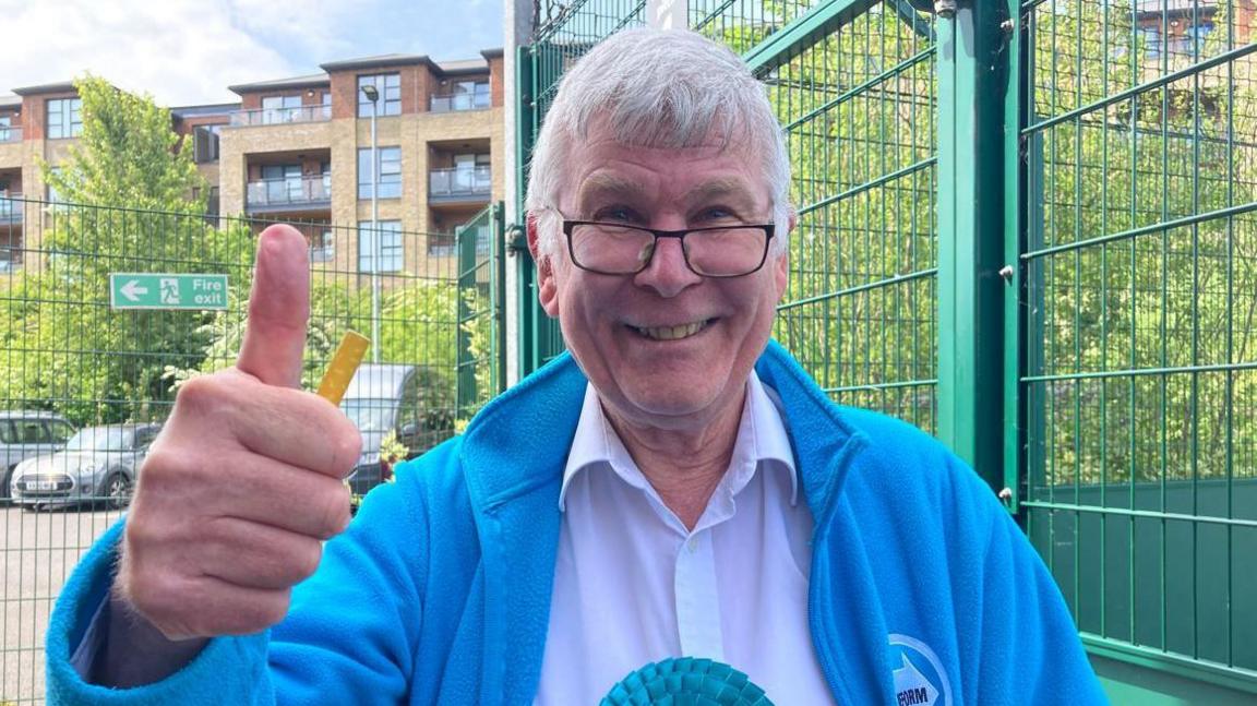 Mr Hopkins has grey hair and is wearing a blue jacket and white shirt with a blue rosette on. He is smiling and giving a thumbs up to the camera. He is stood next to a green metal fence and there is a block of flats behind him.