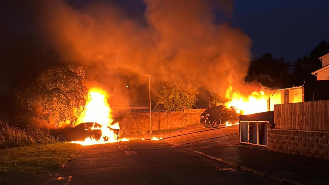 Two cars burn by a wall on a dark street