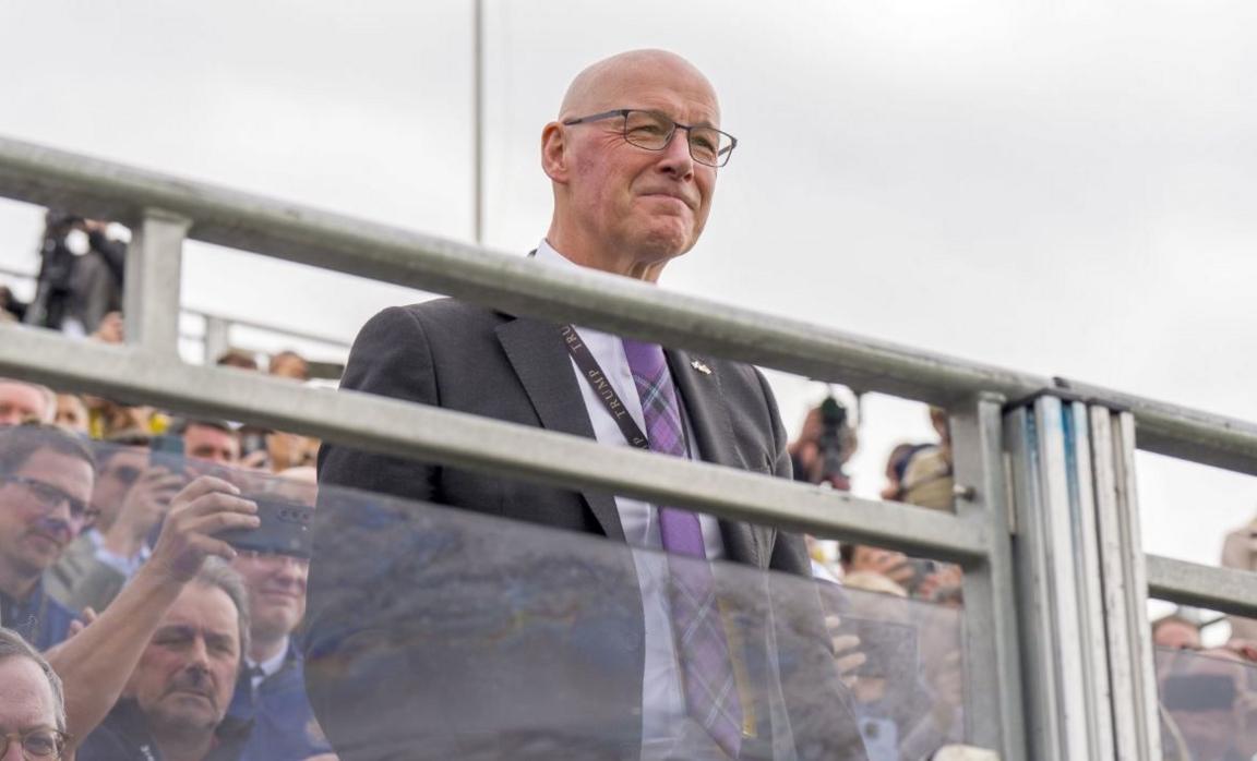 John Swinney with a grey suit and tartan tie stands with a crowd looking over a barrier