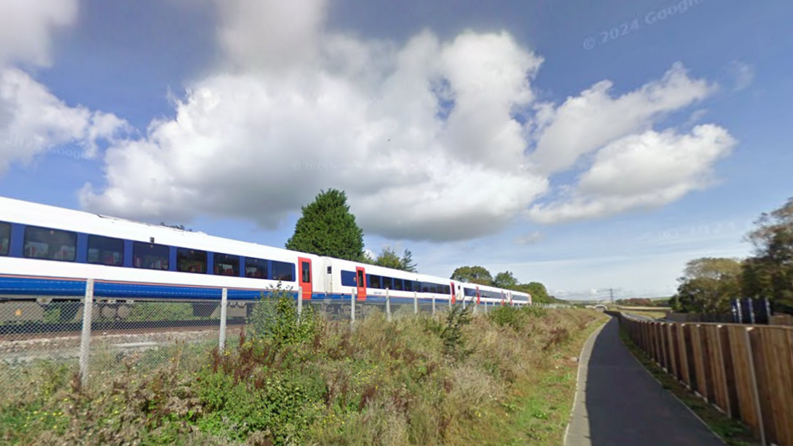 A blue and white train with red doors passes a section of public footpath