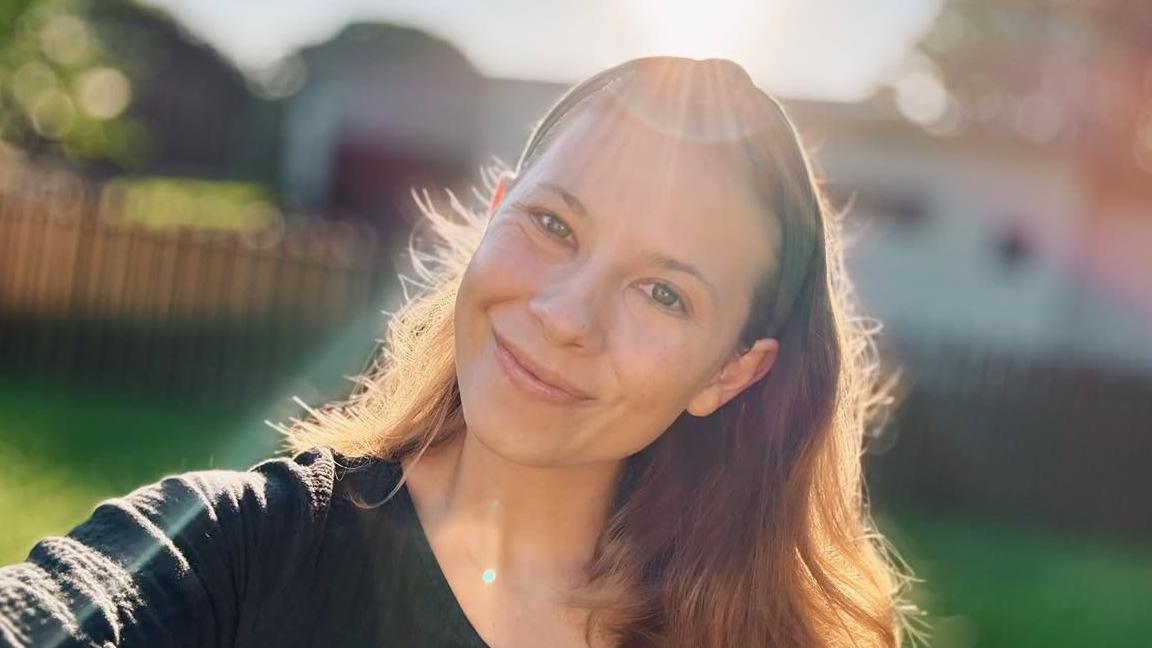 Bindi Irwin, smiling at camera, with shoulder-length brown hair, wearing a black top and sun glare above 